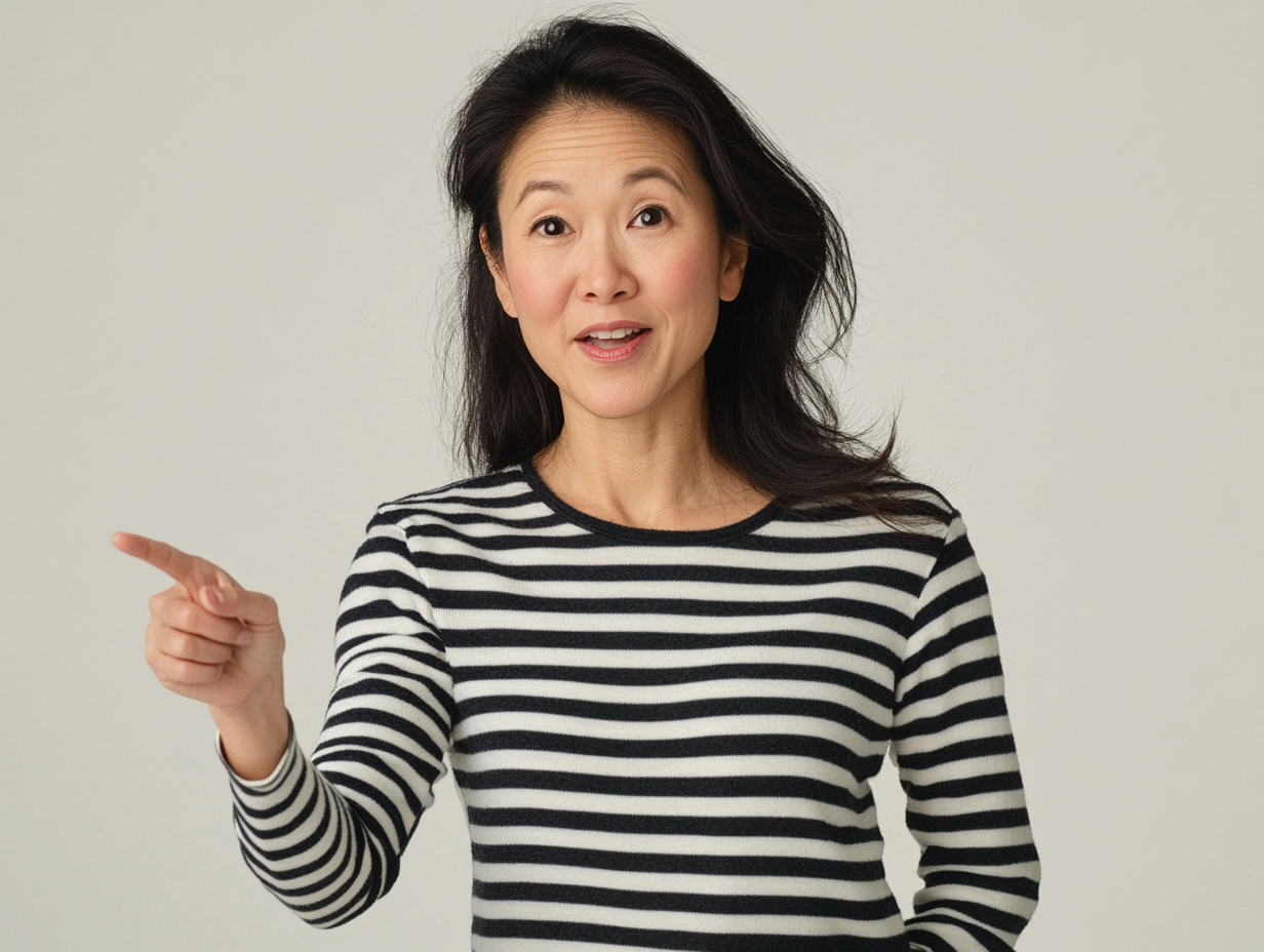 A professional photo of an Asian woman in her late thirties, wearing black and white striped long-sleeved top pointing at something with one finger on plain background.